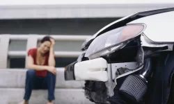 Vrouw zit met handen in het haar naast beschadigde auto.